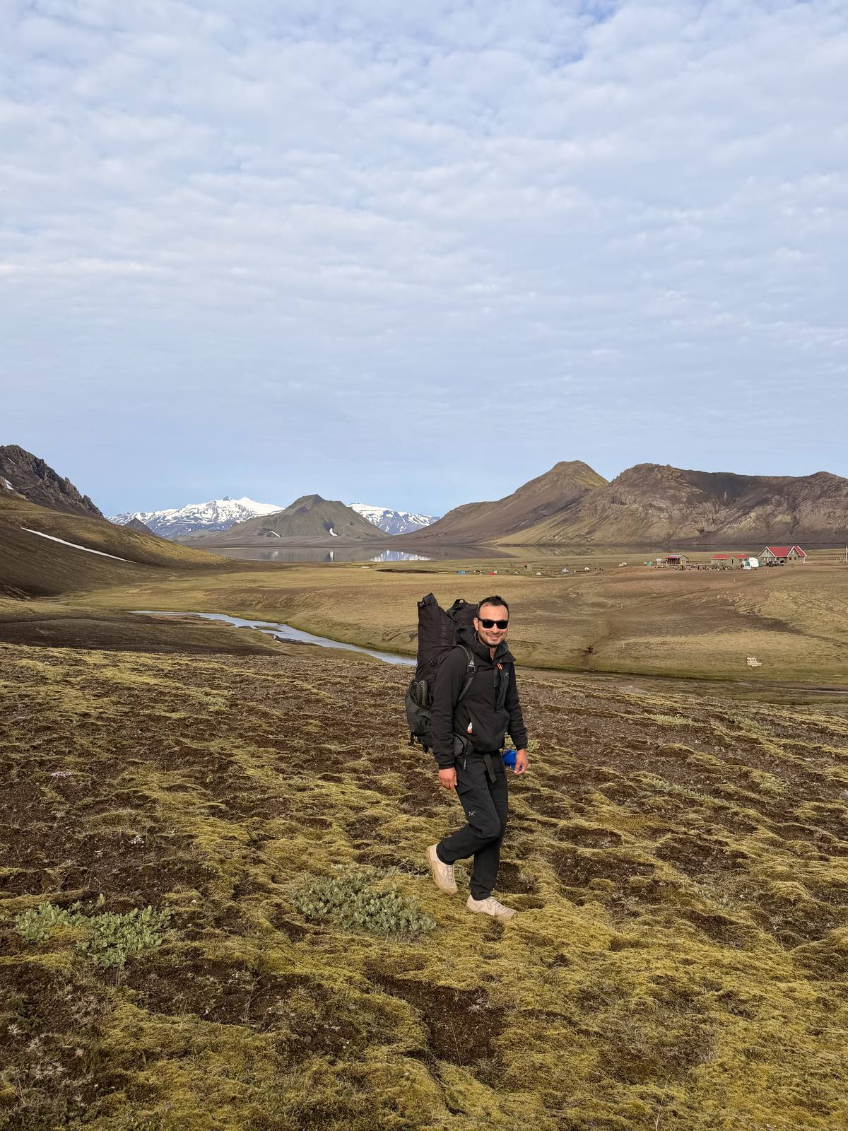 Hiker crossing a mossy valley with mountains and river in the background